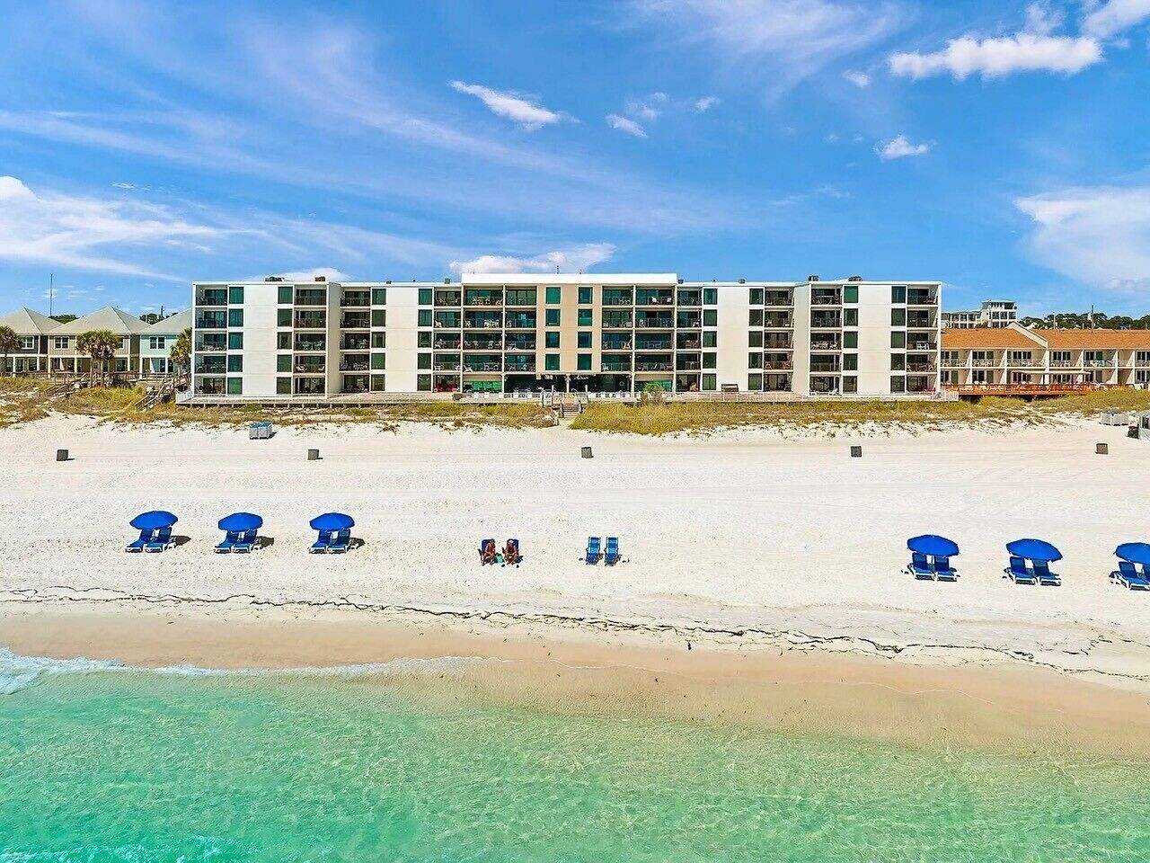 Et hotel ved stranden med flere etager, en sandstrand foran og klart turkis vand.