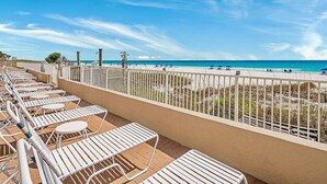 A balcony with white metal chairs and tables, overlooking a beach with people and umbrellas.