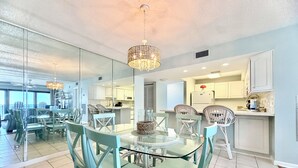 A modern kitchen with white cabinets, a dining area with glass tables and wicker chairs, and a large window offering a view. - Gulfgate 403 - Beautiful Pet Friendly 2 Bed Condo (PANAMA CITY Beach)