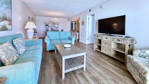 A living room with a sofa, a coffee table, a TV on a wooden stand, and a dining area in the background. - Gulfgate 409 - Pet Friendly Condo on the Beach! (PANAMA CITY Beach)