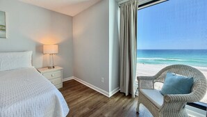 A hotel room with a bed, a nightstand with a lamp, a wicker chair with a cushion, and a view of the beach. - Gulfgate 409 - Pet Friendly Condo on the Beach! (PANAMA CITY Beach)
