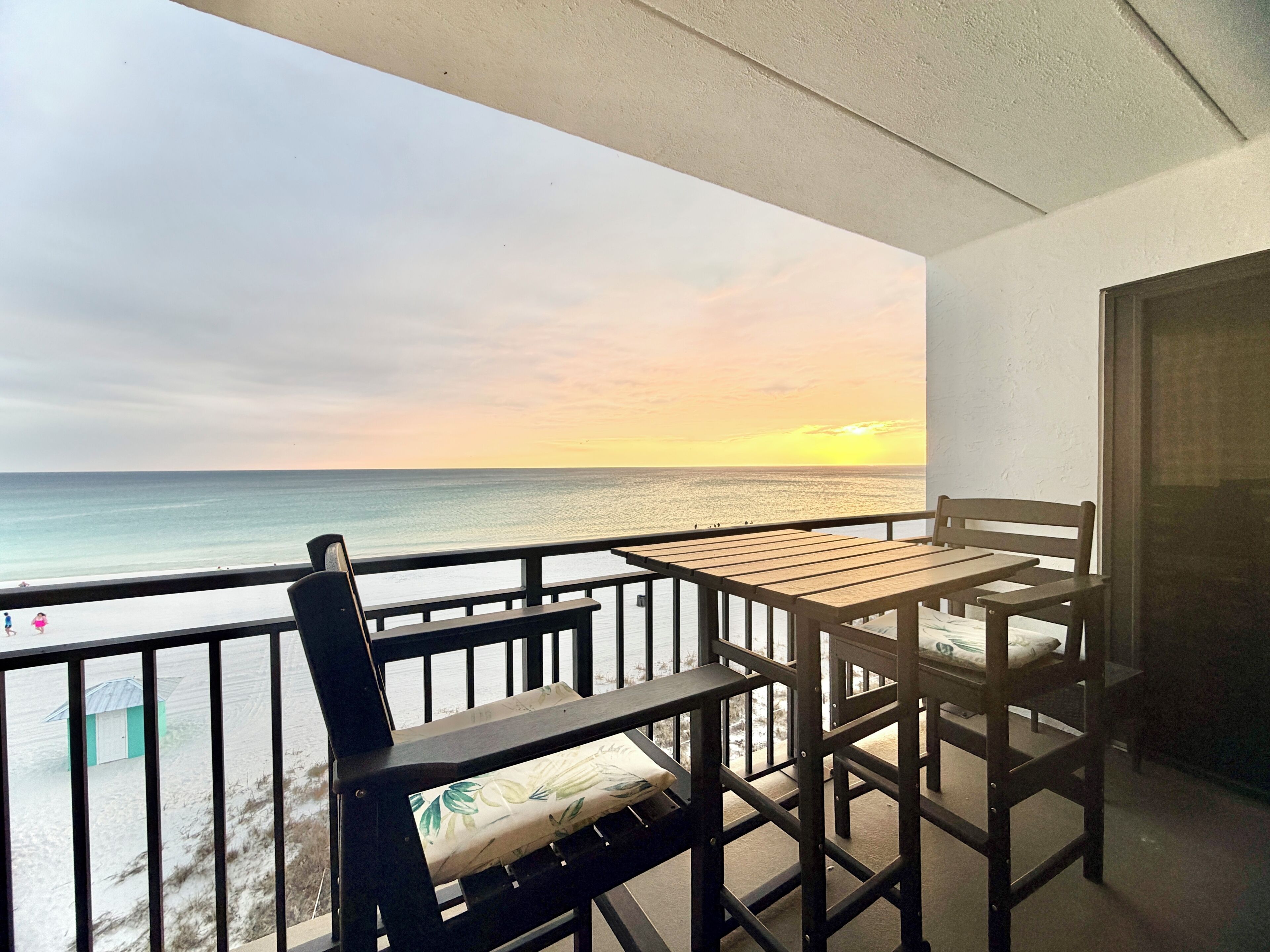 Un balcon avec une table et des chaises en bois, offrant une vue sur l’océan au coucher du soleil.