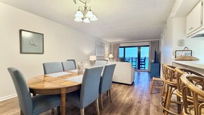 Ein Essbereich mit Holztisch und blauen Stühlen, ein Balkon mit Blick und eine Küchenzeile mit Korbstühlen.