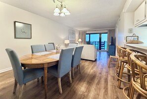 Un coin repas avec une table en bois et des chaises bleues, un balcon avec vue et un espace cuisine avec des chaises en osier.