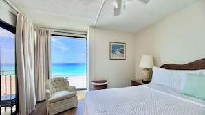 Ein Hotelzimmer mit einem großen Bett, einem Sessel, einem kleinen Tisch, einer Lampe, einem Deckenventilator und Blick auf den Strand.