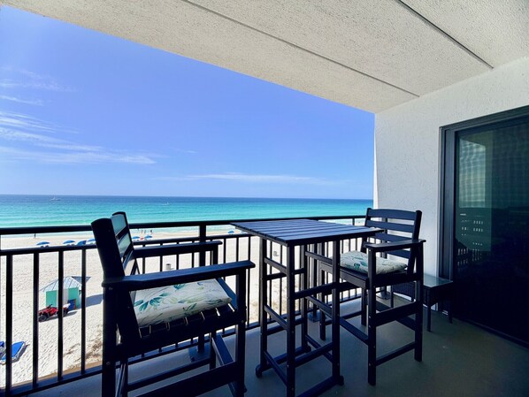A balcony with two chairs and a table, overlooking a beach with umbrellas and a small building.