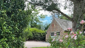 A stone building with a gravel driveway, surrounded by lush greenery and flowering plants. - Court Lodge (Taunton)