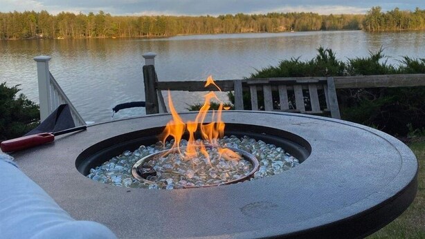 A circular outdoor fire pit with flames, surrounded by a metal railing, overlooking a calm lake with a forested shoreline. - ANNABESSACOOK WATERFRONT WITH DOCK (Winthrop)