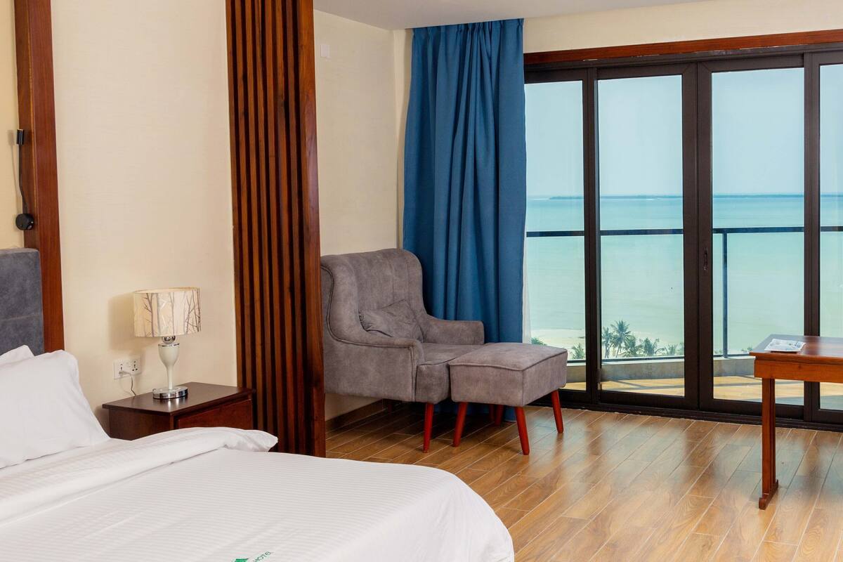 A hotel room with a bed, a chair, a table, a nightstand, and a large window with a view of the sea.