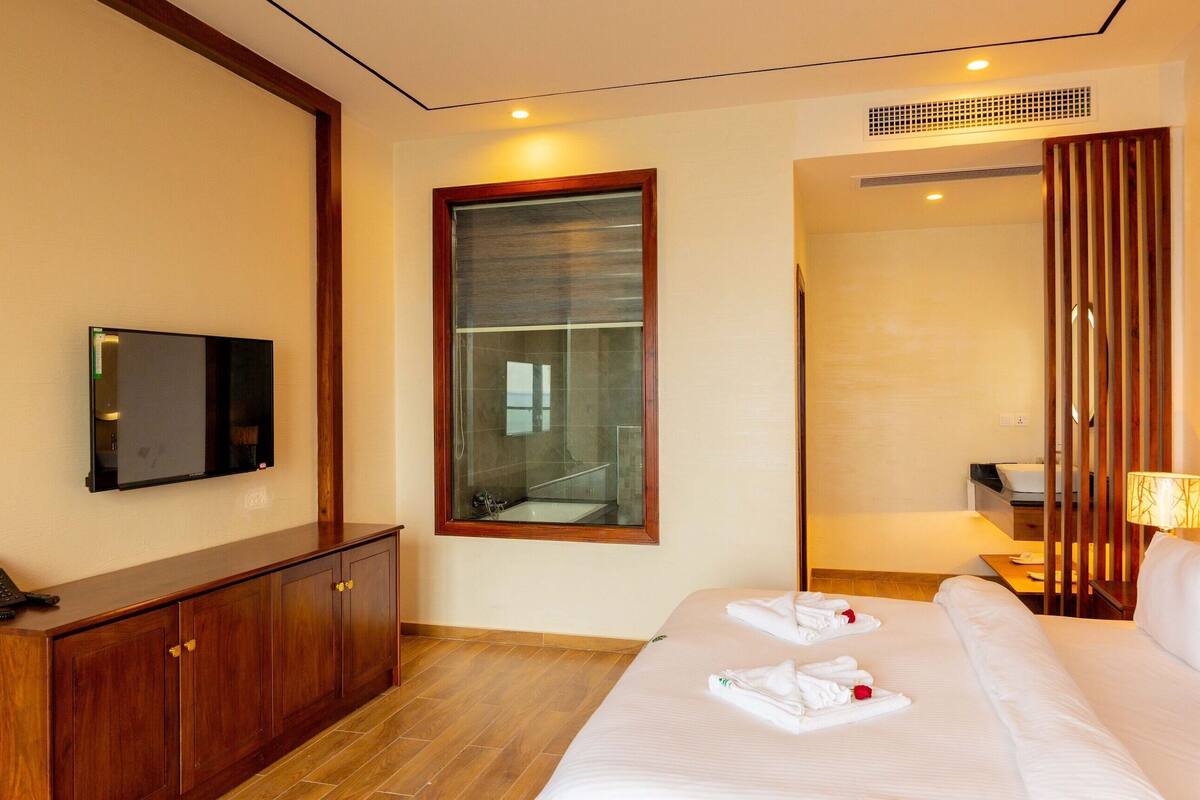 A hotel room with a bed, a TV mounted on the wall, a wooden cabinet, and a bathroom visible through a glass door.