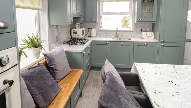 A compact kitchen with teal cabinets, a built-in microwave, a gas stove, and a sink. There is a bench with cushions, a small table, and a potted plant. - Tí Coilín Mhac (Maumeen)