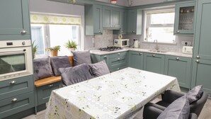 A compact kitchen with teal cabinets, a dining table with a floral tablecloth, and a built-in bench with cushions. - Tí Coilín Mhac (Maumeen)