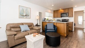 A modern living room with a brown leather sofa, a black ottoman, and a wicker coffee table. A blue chair is positioned near a wooden kitchen island with a microwave and stove. - Harbor House A, overlooking Santa Cruz Harbor (Santa Cruz)