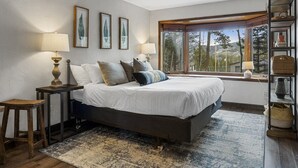 Een slaapkamer met een groot bed, twee nachtkastjes met lampen, een houten kruk, een raam met uitzicht en een boekenkast.
