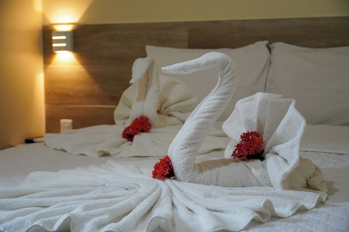 A hotel room with a neatly made bed featuring swan-shaped towels and red flowers.