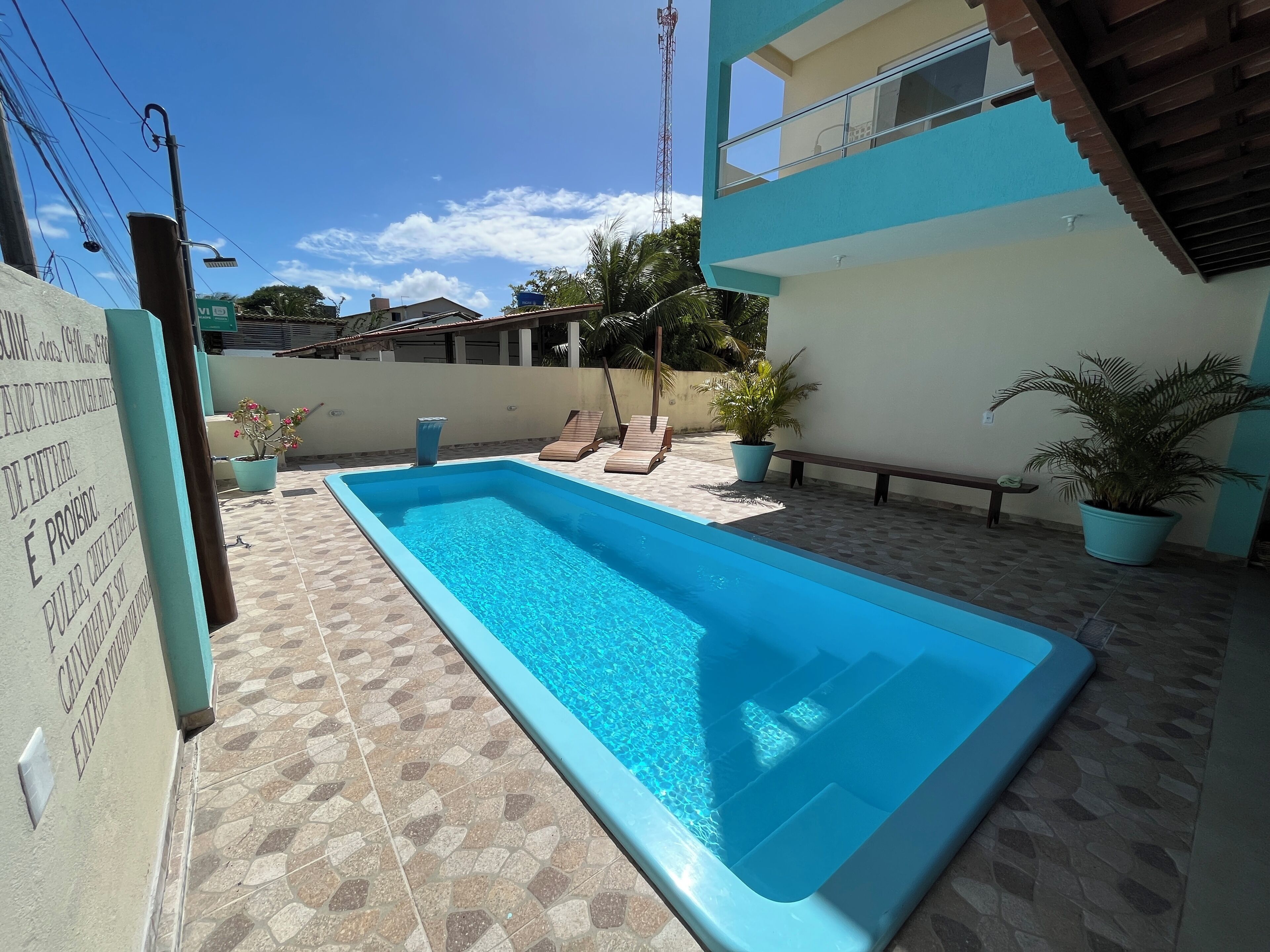 Piscina retangular com deck ladrilhado, rodeada por plantas em vasos e espreguiçadeiras.