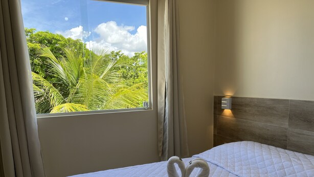 A hotel room with a bed, a window with curtains, and a view of greenery outside. - Pousada Donna Maraka (Ipojuca)