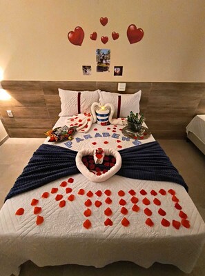 Una cama con un arreglo de pétalos de rosa en forma de corazón, una manta con el nombre "RABEN" y una mesita de noche con un cojín con forma de corazón.