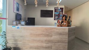 A reception desk with a light wood finish, a computer monitor, and a telephone. There are informational posters on the wall and a potted plant to the left. - Pousada Donna Maraka (Ipojuca)