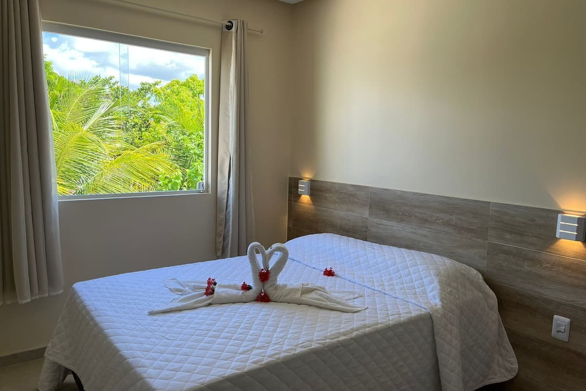 A hotel room with a bed, curtains, a window with a view of greenery, and wall-mounted lights.