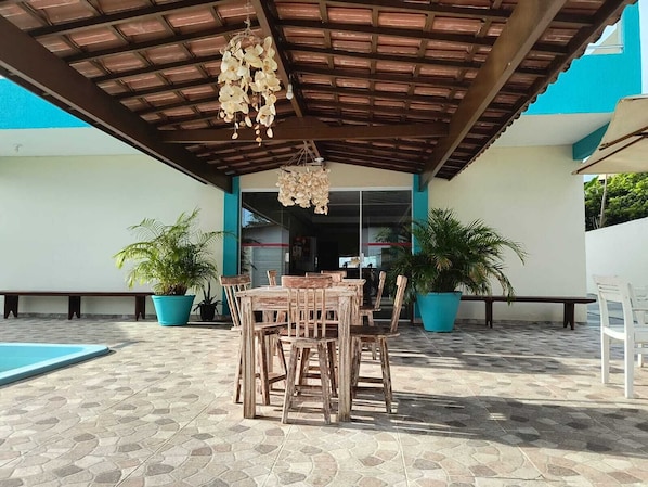 An outdoor patio area with a wooden table and chairs, potted plants, and a pool. - Pousada Donna Maraka (Ipojuca)