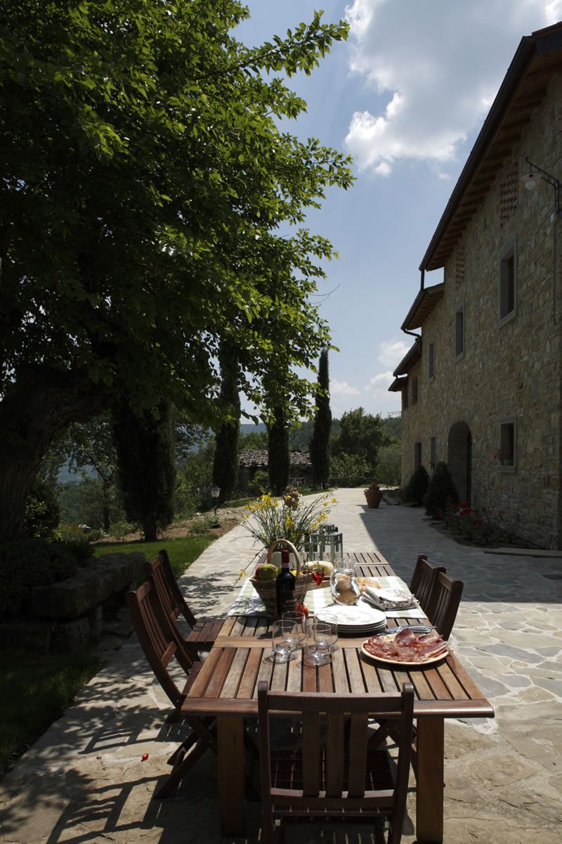 An outdoor dining area with a stone table and chairs, surrounded by greenery and a stone building.