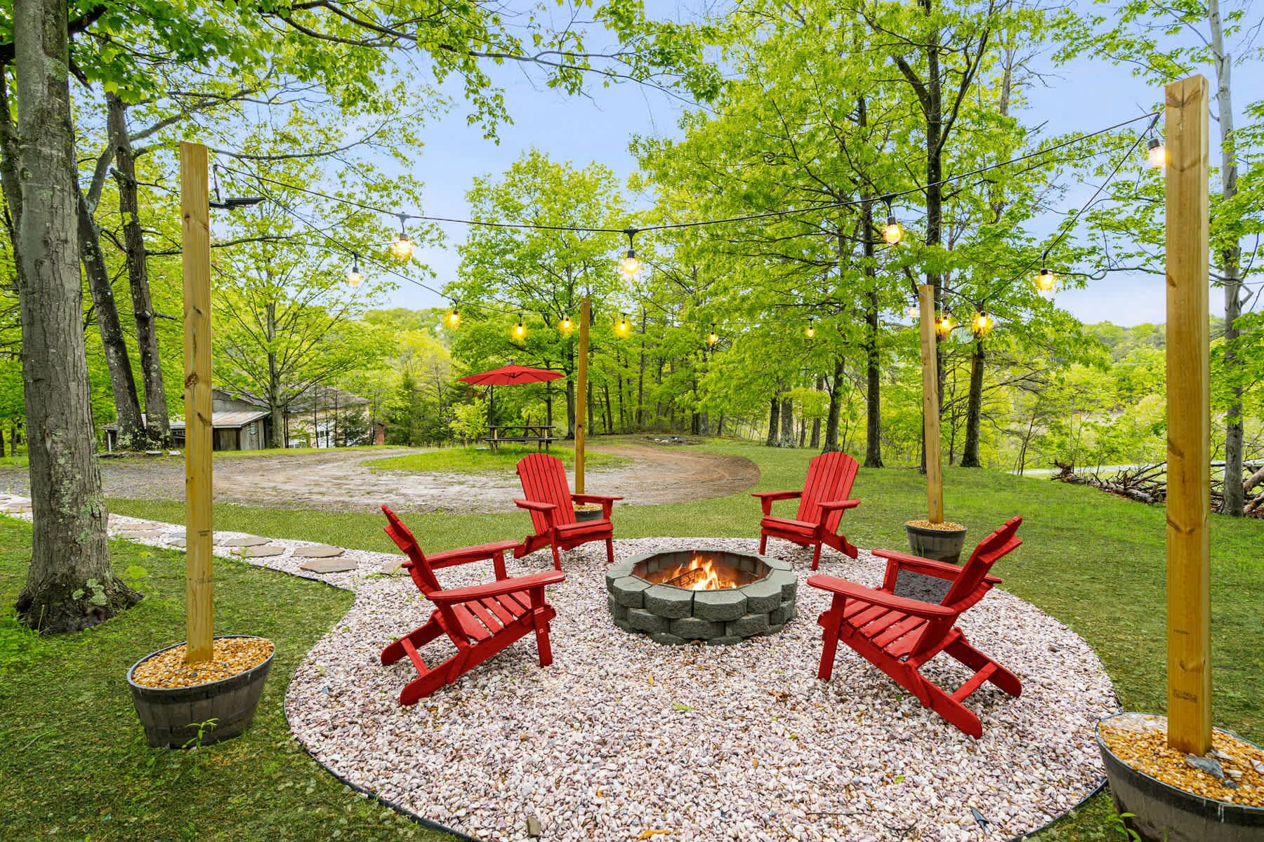 An outdoor seating area with a fire pit, surrounded by red Adirondack chairs, and string lights overhead.