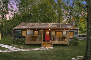 A log cabin with a red door, a porch with string lights, and a stone pathway leading to the entrance. - Modernized Log Cabin w Hot Tub, Fire Pit & Views! (Berkeley Springs)