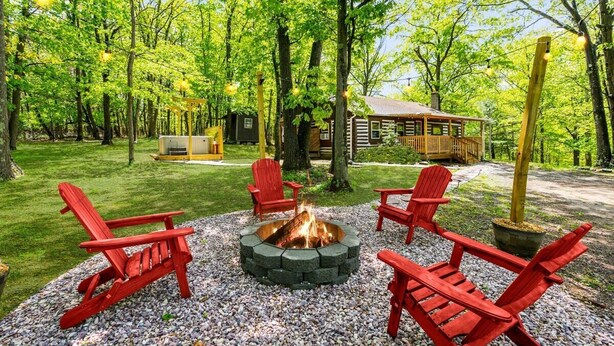 A forested campsite with a fire pit, red Adirondack chairs, and a cabin. - Modernized Log Cabin w Hot Tub, Fire Pit & Views! (Berkeley Springs)