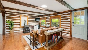 A log cabin interior with a wooden floor, a sofa, a table, a fireplace, and a door with windows. - Modernized Log Cabin w Hot Tub, Fire Pit & Views! (Berkeley Springs)