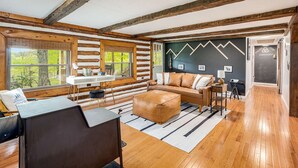 A log cabin interior with a wooden floor, a sofa, a desk, and a view of the outdoors. - Modernized Log Cabin w Hot Tub, Fire Pit & Views! (Berkeley Springs)