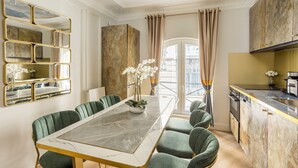 Un coin repas avec une table en marbre et des chaises recouvertes d’un tissu vert, un miroir décoratif et, à travers une porte vitrée, une vue sur un balcon extérieur.