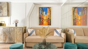Un salon moderne avec un canapé beige, des poufs turquoise, une table basse en verre et deux tableaux aux couleurs vives représentant des paysages urbains, accrochés aux murs.