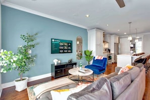 A modern living room with a blue accent wall, a flat-screen TV, a potted plant, a sofa, a coffee table, and a kitchen in the background. - Summer game room, chef’s kitchen, garage parking near Broadway & Gulch (Nashville)