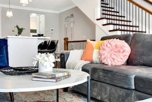 A modern living room with a grey sofa, a white coffee table with books and a vase of flowers, a staircase with wooden railings, and a kitchen area in the background. - Summer game room, chef’s kitchen, garage parking near Broadway & Gulch (Nashville)