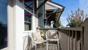 Une terrasse en bois avec deux chaises blanches et une petite table, une fenêtre ornée d’une enseigne au néon et un mur de briques.