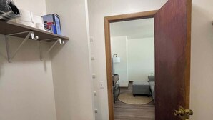 A hallway with a wooden door, a shelf with toiletries, and a glimpse of a living room with a sofa and a lamp.