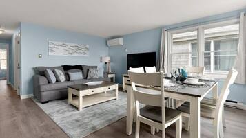 Een moderne woonkamer met een grijze bank, een bijzettafeltje en een eethoek met een witte tafel en stoelen.