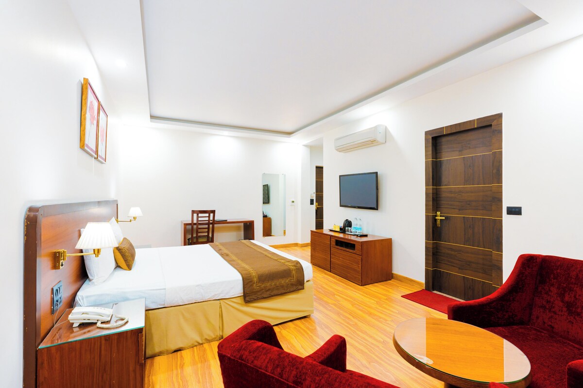 A hotel room with a bed, desk, chair, TV, and a red velvet armchair.