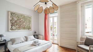 Ein Schlafzimmer mit einem Bett, einem Nachttisch, einer Lampe, einem großen Gemälde, einem Korbstuhl und einer Glastür zu einem Balkon.