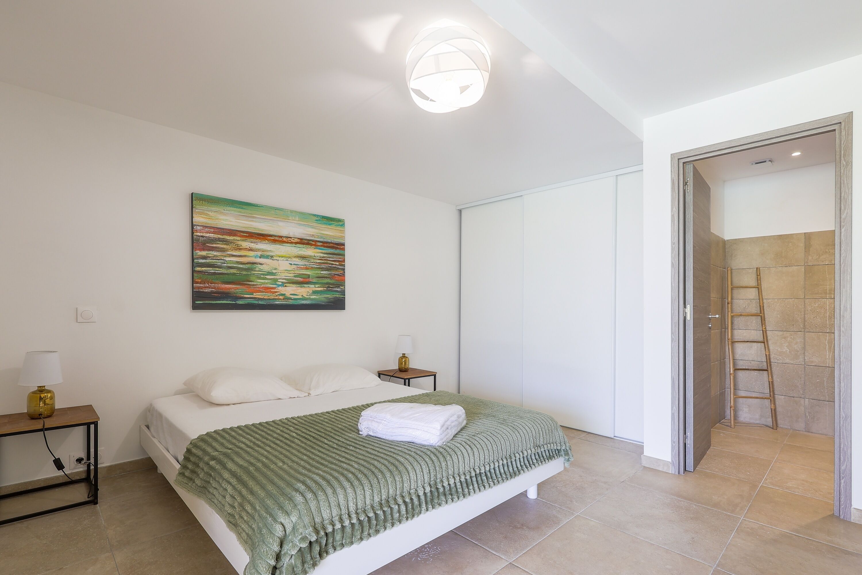 A bedroom with a bed, a painting, a nightstand, and a door leading to a bathroom.