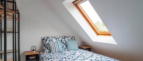 A bedroom with a skylight, a bed with patterned bedding, a bedside table with a lamp, and a wooden ladder.