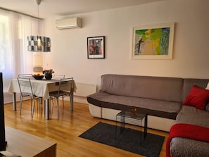 A living room with a sofa, a dining table with chairs, a small side table, and a painting on the wall.