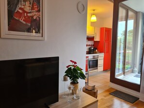 A modern living room with a flat-screen TV, a potted plant, and a dining area with red cabinets and a kitchen in the background.