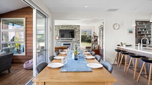 Un espace de repas moderne avec une longue table dressée pour six personnes, une cheminée en pierre et une cuisine visible à travers une porte ouverte.