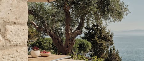 A stone wall, a wicker chair, a table with a bowl of fruit, and a view of the sea.