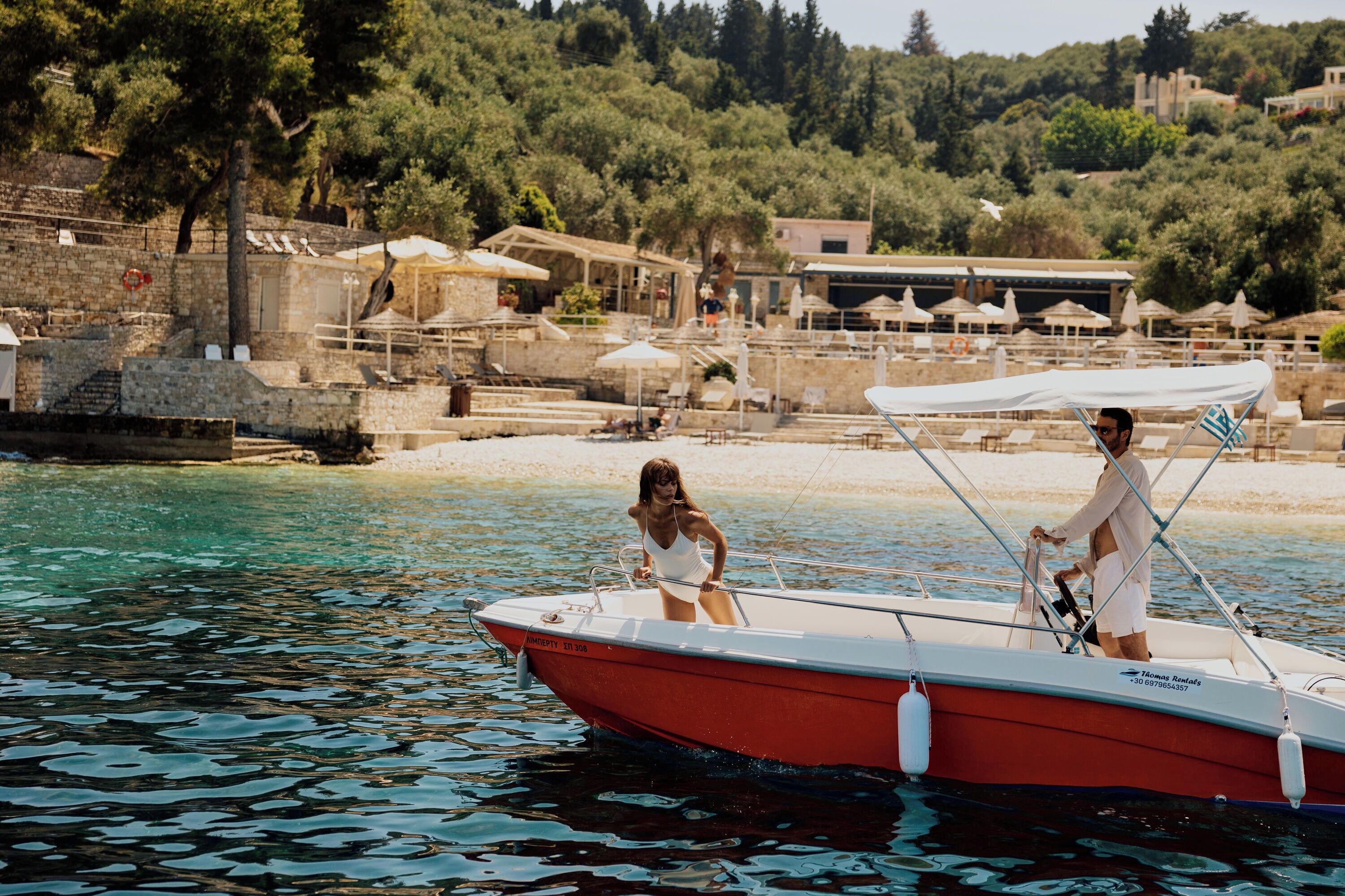 A red boat with two people on it, docked near a beach with buildings and trees in the background.