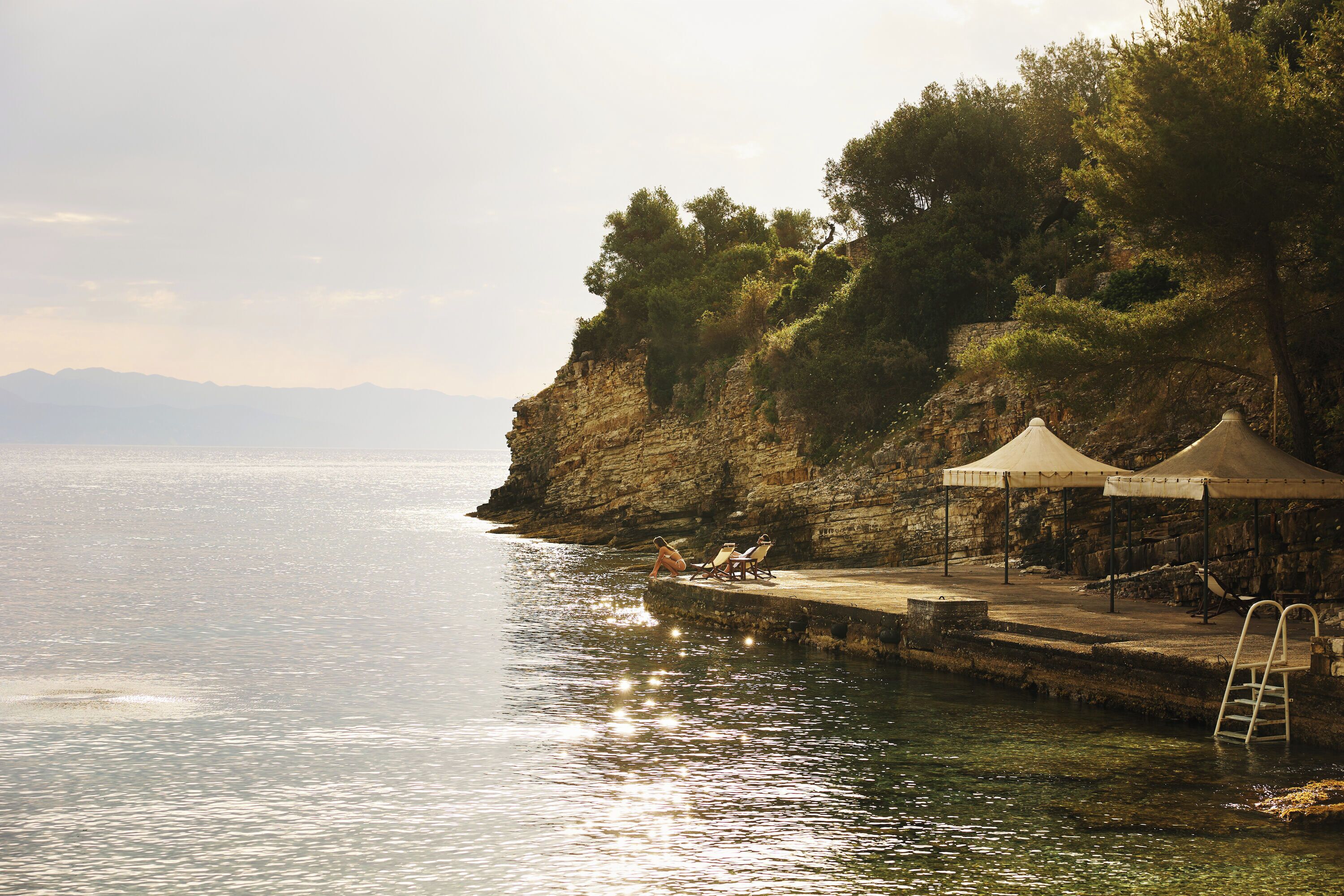 A coastal scene with a pier extending into calm waters, a rocky cliff with trees, and a clear sky.