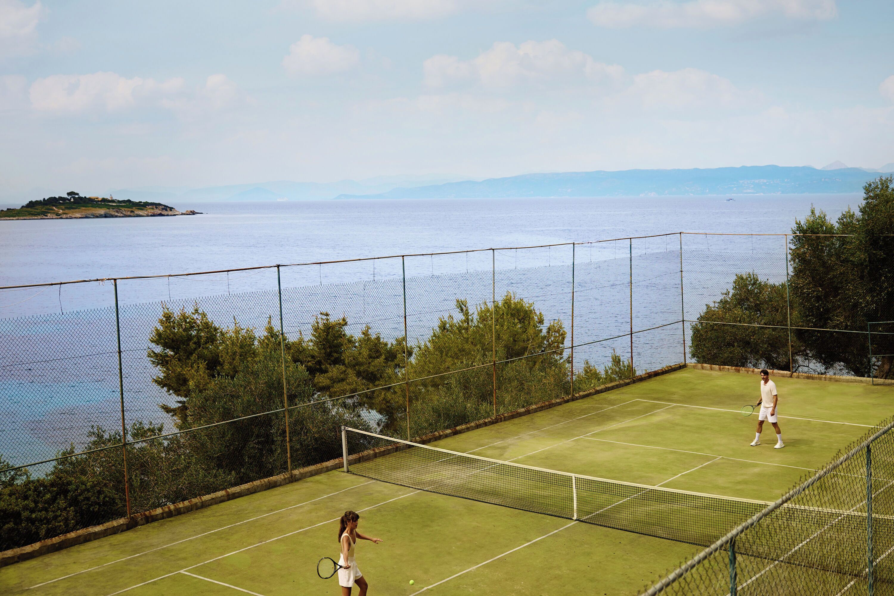 Un court de tennis avec un filet, entouré d’une clôture, et offrant une vue sur une étendue d’eau et des collines lointaines.