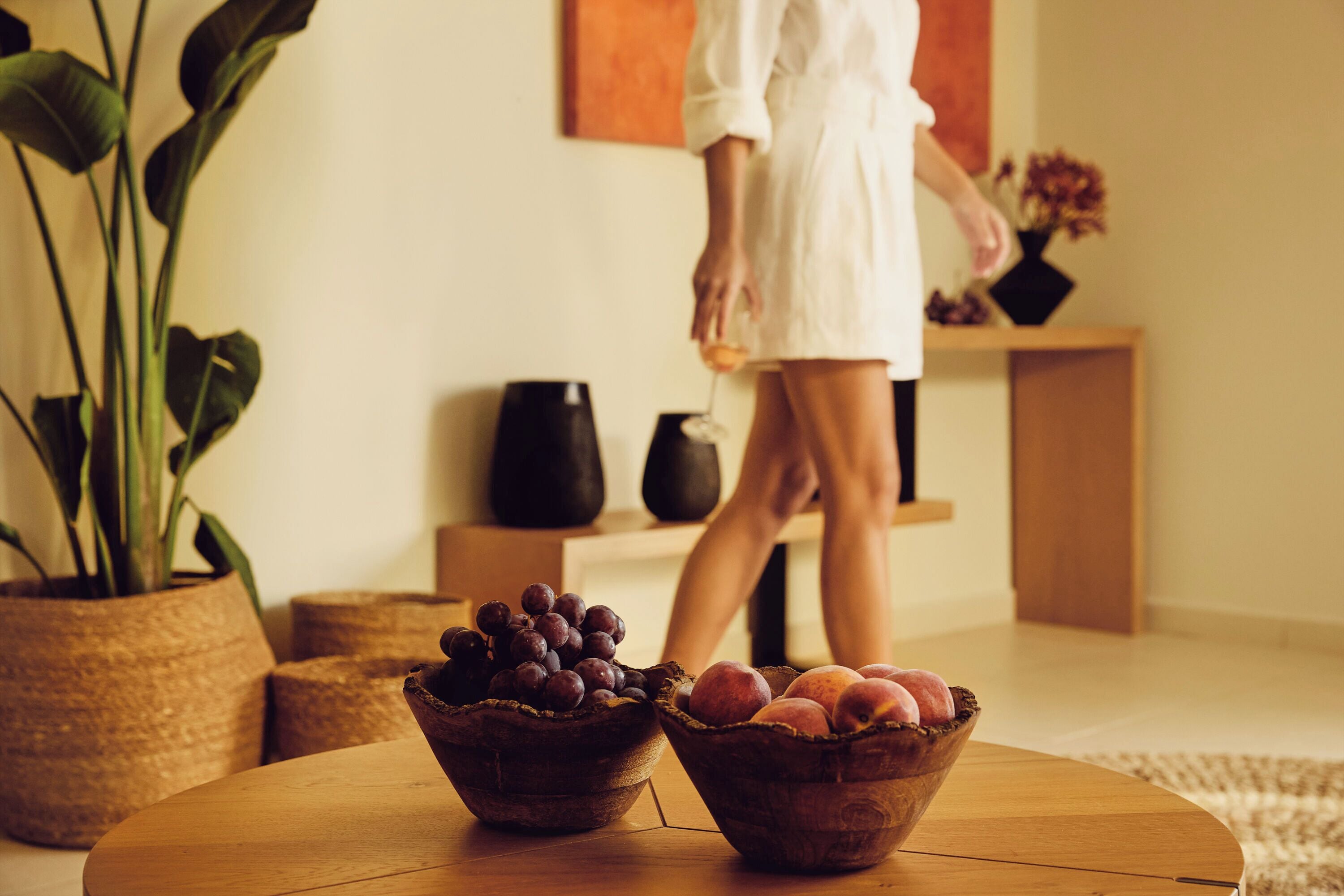 A room with a round wooden table holding two woven baskets, one with grapes and the other with peaches. There is a potted plant to the left and a vase with flowers on a shelf in the background.